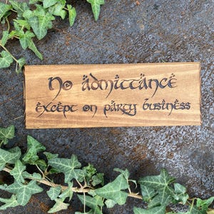 Può includere: Un cartello rettangolare in legno con la scritta "No admittance except on party business" in un carattere stilizzato. Il cartello è circondato da edera verde su una superficie scura e strutturata.