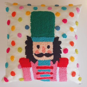Puede incluir: Una almohada blanca con un diseño de cascanueces bordado de colores. El cascanueces lleva un sombrero verde, una chaqueta rosa y una faja azul. El fondo de la almohada es blanco con lunares de colores.