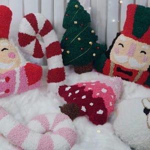 Puede incluir: Una colección de cojines decorativos con temática navideña. Los cojines presentan diseños como Papá Noel, un cascanueces, un árbol de Navidad y bastones de caramelo. Están hechos de un material suave y texturizado en colores festivos como rojo, rosa, verde y blanco.