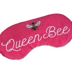 以下が含まれることがあります： ピンクのアイマスクに、白い刺繍で「Queen Bee」と刺繍された小さなミツバチが、黒と黄色のストライプで刺繍されています。