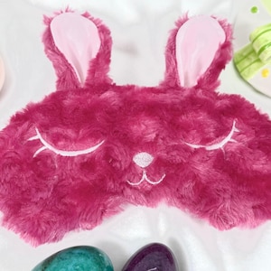 May include: A pink fuzzy sleep mask with bunny ears. The mask has embroidered white eyes and a nose.