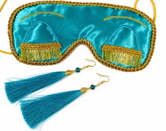 May include: A turquoise satin sleep mask featuring gold embroidered eyelashes and a gold braided trim, accompanied by a pair of matching turquoise tassel earrings with gold-tone hardware and small dark beads on the earring hooks.