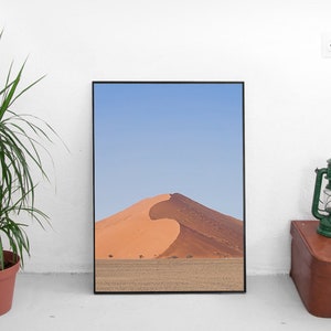 Può includere: Una fotografia incorniciata di una grande duna di sabbia arancione in un paesaggio desertico. Il cielo è blu e la sabbia è di un colore arancione caldo.