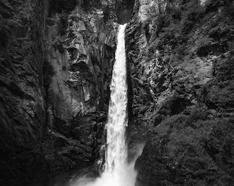 May include: A black and white photograph of a waterfall cascading down a rocky cliff face. The water is flowing over the edge of the cliff and into a pool below. The surrounding area is covered in trees and vegetation.