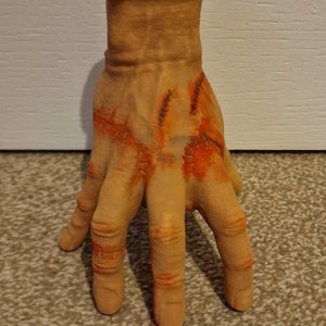 Op de afbeelding: Een realistisch ogende, gebeeldhouwde hand met gestikte wonden en oranje-rode accenten. De hand is gemaakt van lichtbruin materiaal en is geposeerd met gespreide vingers.