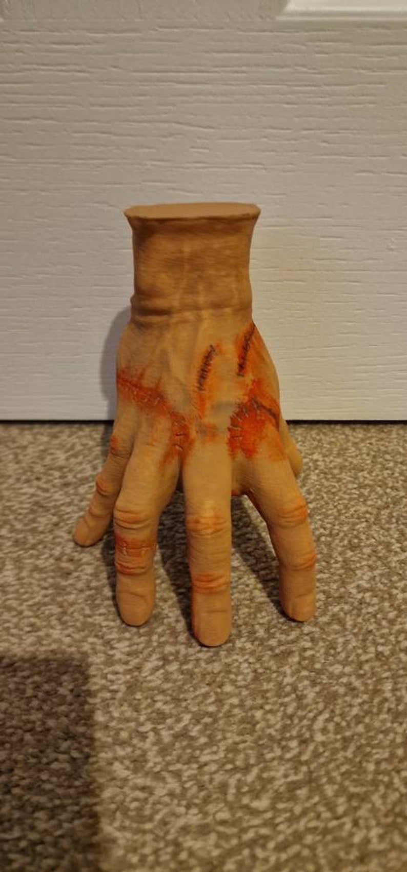 Op de afbeelding: Een realistisch ogende, gebeeldhouwde hand met gestikte wonden en oranje-rode accenten. De hand is gemaakt van lichtbruin materiaal en is geposeerd met gespreide vingers.