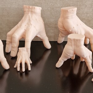 Op de afbeelding: Zes verschillende formaten, 3D-geprinte, vleeskleurige handen, staand op een zwart oppervlak. De handen staan allemaal in verschillende poses, met sommige vingers gestrekt en andere gekruld.