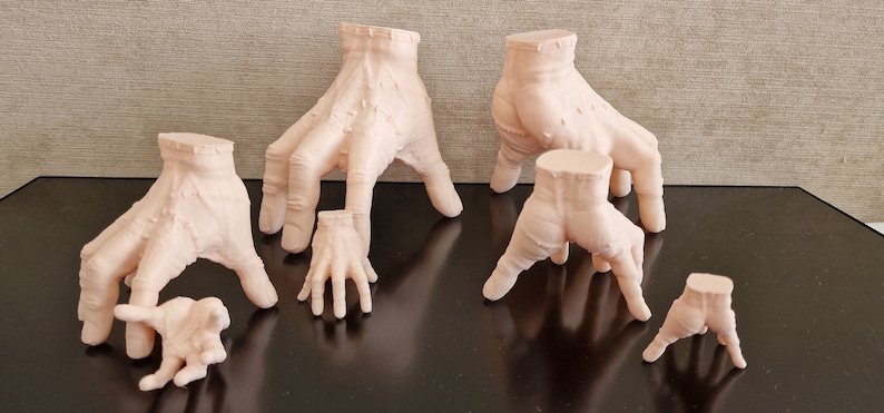 Op de afbeelding: Zes verschillende formaten, 3D-geprinte, vleeskleurige handen, staand op een zwart oppervlak. De handen staan allemaal in verschillende poses, met sommige vingers gestrekt en andere gekruld.