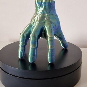 Op de afbeelding: Een 3D-geprint handbeeld met een groene en blauwe iriserende afwerking. De hand is geposeerd met gestrekte vingers en rust op een zwarte ronde basis.