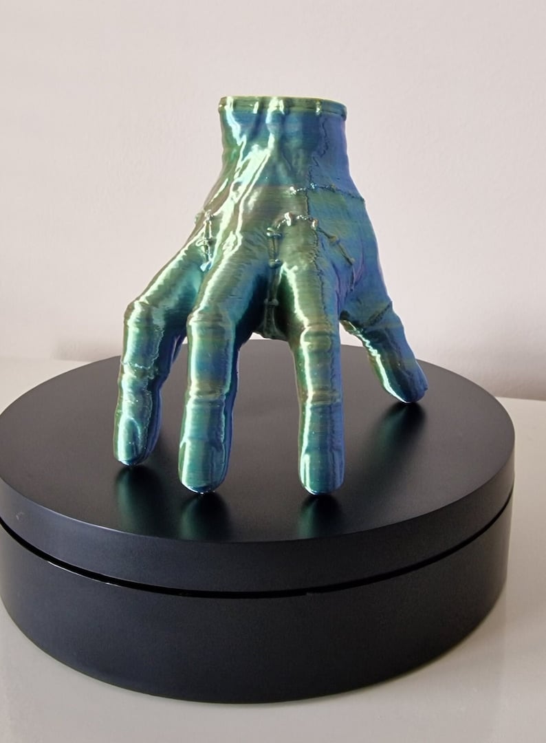 Op de afbeelding: Een 3D-geprint handbeeld met een groene en blauwe iriserende afwerking. De hand is geposeerd met gestrekte vingers en rust op een zwarte ronde basis.