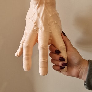 Op de afbeelding: Een lichtroze keramische hand met gestikte naden, die lijkt op een monsterhand, wordt vastgehouden door een persoon met donkerrode nagellak.
