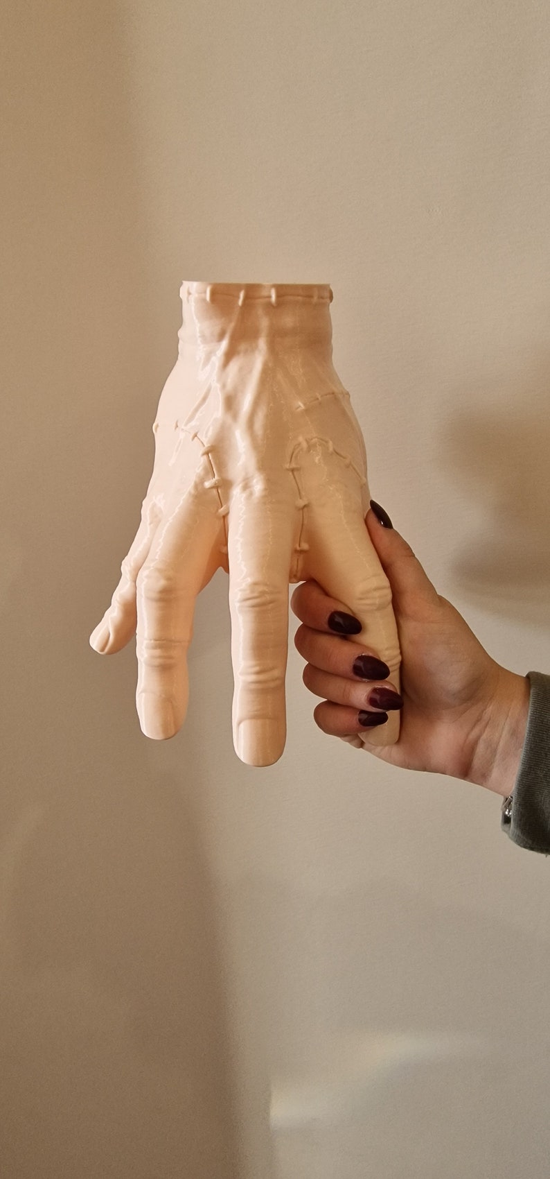Op de afbeelding: Een lichtroze keramische hand met gestikte naden, die lijkt op een monsterhand, wordt vastgehouden door een persoon met donkerrode nagellak.