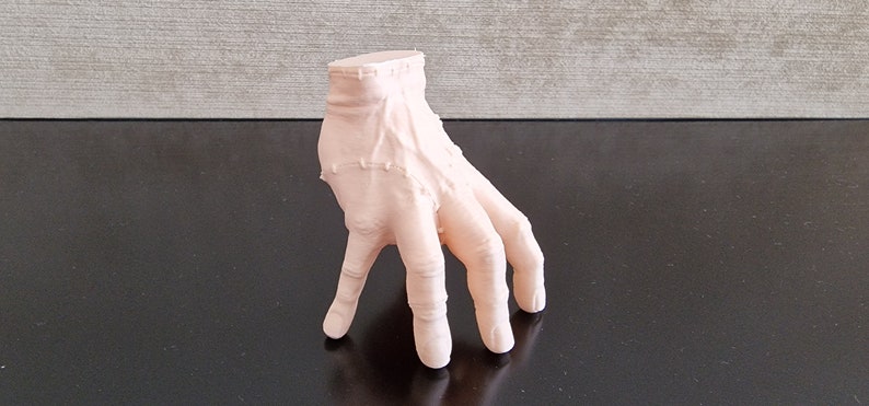 Op de afbeelding: Een 3D-geprint model van een menselijke hand met alle vingers gestrekt. De hand is gemaakt van lichtroze plastic en is geposeerd alsof hij reikt.