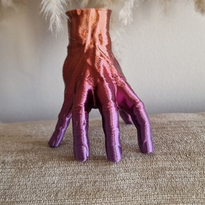 Op de afbeelding: Een handvormige vaas met een kleurverloop van oranje naar paars. De vaas is gemaakt van een glad, glanzend materiaal en heeft een uniek, abstract ontwerp.