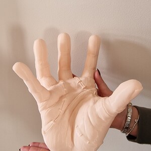 Op de afbeelding: Een groot, lichtroze, gedetailleerd, realistisch handbeeld met gestikte naden. De hand wordt vastgehouden door een hand van een persoon.
