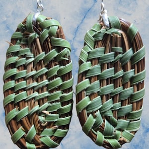 May include: A pair of handmade earrings featuring an oval woven design. The earrings are crafted from natural brown and green materials, with a woven pattern. The earrings have silver-colored ear wires.