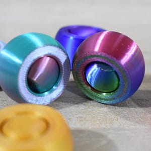 May include: Five colorful, round, 3D printed fidget toys. The toys are made of smooth plastic and have a spinning top design. The colors are silver, teal, purple, pink, and gold.