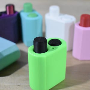 May include: A set of six colorful cases for small electronic devices. The cases are made of a soft, flexible material and come in a variety of colors, including purple, teal, white, pink, and green. Each case has a unique design and features a small button on the top.