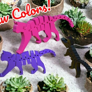 May include: Three 3D printed articulated cat figurines in pink, purple, and black. The cats are posed in a walking position. The figurines are displayed on a white surface with small potted plants.