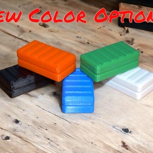 May include: Five small rectangular blocks in different colors: orange, green, white, blue, and black. The blocks are stacked on top of each other, with the orange block on top of the black block. The blocks are made of a smooth, textured material.  The text "NEW COLOR OPTIONS!" is written in red letters across the top of the image.