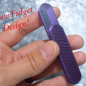 May include: A purple fidget tool with a textured surface on one side and a smooth surface on the other side. The text "New Fidget Design!" is visible in the image.