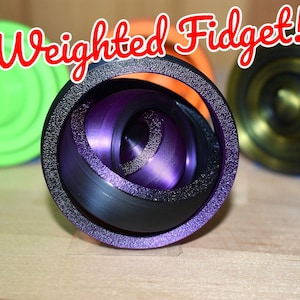 May include: A collection of weighted fidget toys in various colors. The central fidget is a deep purple and black design, with a textured outer ring. Other fidgets are green and gold. The text "Weighted Fidget!" is in red.