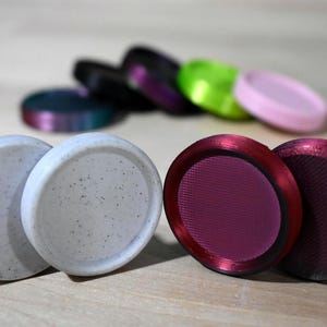 May include: A collection of colorful, round objects. Two light gray, speckled discs are in the foreground. Behind them are other discs in shades of red, purple, green, pink, and black. The objects appear to be coasters or decorative items.