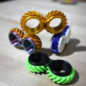 May include: Four fidget toys in different colors. The top toy is gold and has a gear design. The other three toys are purple, blue, and green. All of the toys have a gear design.
