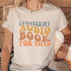 May include: A beige t-shirt with the text "I paused my audiobook for this" in a retro font with colorful letters.