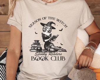 May include: A beige t-shirt with a black and white illustration of a witch reading a book. The text on the shirt reads "Season of the Witch Spooky Readers Book Club".