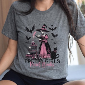 May include: Gray t-shirt featuring a witch reading a book, surrounded by bats and a black cat. The witch wears a black hat and pink dress. The text "PRETTY GIRLS Read Books" is printed below the illustration.