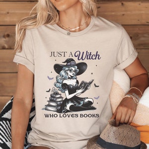Puede incluir: Una camiseta beige con un gráfico de una bruja leyendo un libro. El texto en la camiseta dice "Just a Witch who loves books".