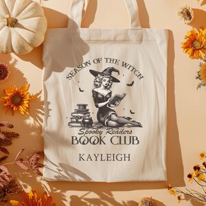 Puede incluir: Una bolsa de tela blanca con una ilustración en blanco y negro de una bruja leyendo un libro. El texto en la bolsa dice "Season of the Witch Spooky Readers Book Club KAYLEIGH".