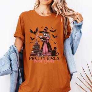 May include: Orange t-shirt with a graphic of a witch reading a book, surrounded by bats and a black cat. The text "Pretty Girls Read Books" is below the image. The shirt is a casual style.