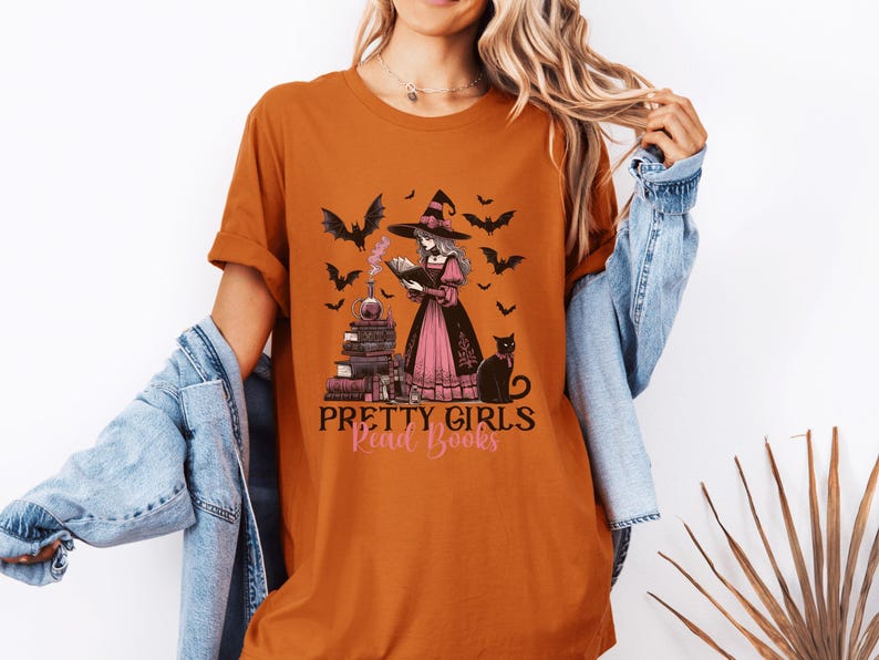 May include: Orange t-shirt with a graphic of a witch reading a book, surrounded by bats and a black cat. The text "Pretty Girls Read Books" is below the image. The shirt is a casual style.