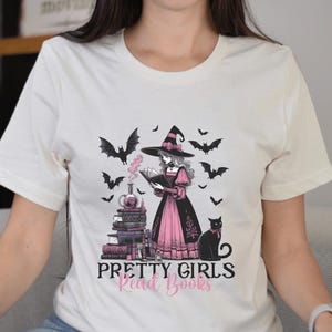 May include: Cream-colored t-shirt featuring a graphic of a witch reading a book, surrounded by bats and a black cat. The witch is wearing a black hat and pink dress. Text on the shirt reads "Pretty Girls Read Books."