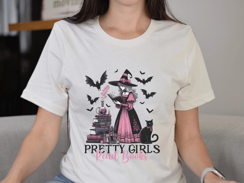 May include: Cream-colored t-shirt featuring a graphic of a witch reading a book, surrounded by bats and a black cat. The witch is wearing a black hat and pink dress. Text on the shirt reads "Pretty Girls Read Books."