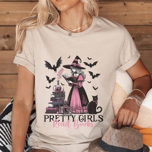 May include: Beige t-shirt with a graphic of a witch reading a book, surrounded by bats and a black cat. The text "Pretty Girls Read Books" is below the illustration.