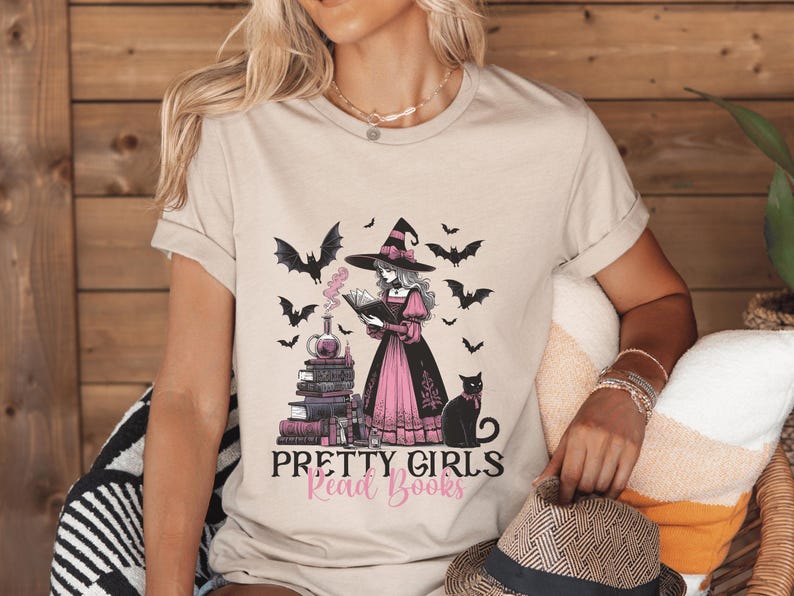 May include: Beige t-shirt with a graphic of a witch reading a book, surrounded by bats and a black cat. The text "Pretty Girls Read Books" is below the illustration.