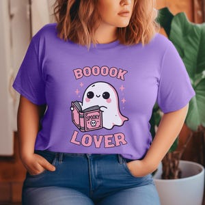 May include: A lilac-colored t-shirt featuring a cute ghost reading a book with the word "Spooky" on the cover. The text "BOOOOK LOVER" is printed above and below the ghost illustration in pink. The shirt is a casual wear item.