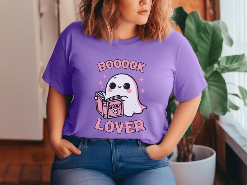 May include: A lilac-colored t-shirt featuring a cute ghost reading a book with the word "Spooky" on the cover. The text "BOOOOK LOVER" is printed above and below the ghost illustration in pink. The shirt is a casual wear item.