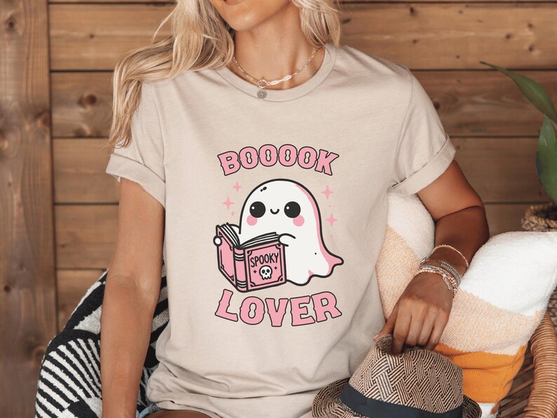 May include: Beige t-shirt featuring a cartoon ghost reading a pink book with the words "Spooky" and a skull on the cover. The text "BOOOK LOVER" is above and below the ghost illustration. The shirt has rolled-up sleeves.