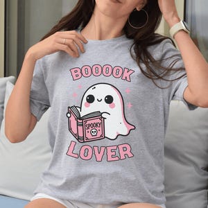 May include: A gray t-shirt featuring a cartoon ghost reading a book. The ghost is white with black eyes and a pink blush. The text "BOOOOK LOVER" is printed in pink, with stars. The book says "Spooky".