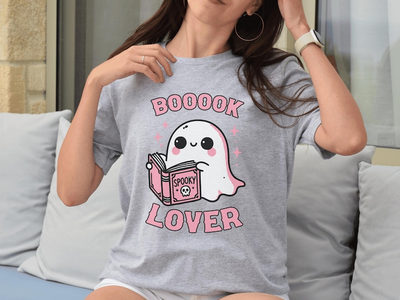 May include: A gray t-shirt featuring a cartoon ghost reading a book. The ghost is white with black eyes and a pink blush. The text "BOOOOK LOVER" is printed in pink, with stars. The book says "Spooky".