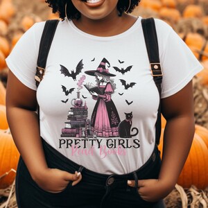 May include: White t-shirt featuring a witch reading a book, surrounded by bats and a black cat. The witch wears a black hat and pink dress. The text "PRETTY GIRLS Read Books" is printed below the image.