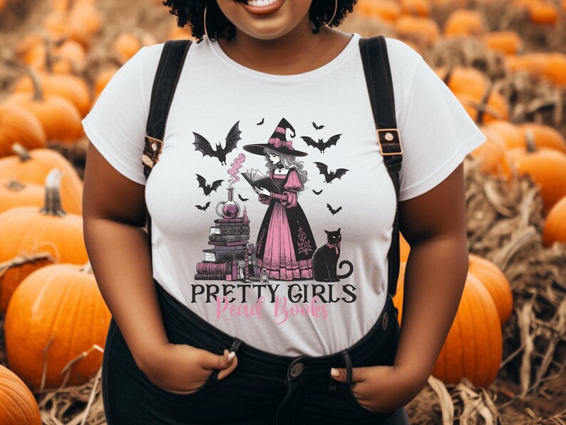 May include: White t-shirt featuring a witch reading a book, surrounded by bats and a black cat. The witch wears a black hat and pink dress. The text "PRETTY GIRLS Read Books" is printed below the image.
