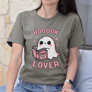 May include: Olive green t-shirt featuring a cute cartoon ghost reading a pink book with the words "BOOOOK LOVER" in pink. The ghost has a smiling face and is surrounded by pink stars. A fun, spooky design.