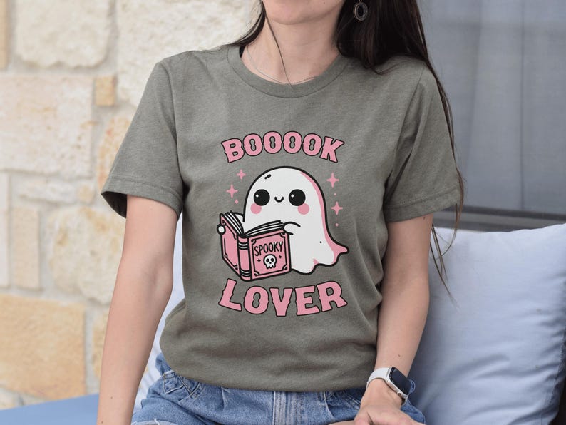 May include: Olive green t-shirt featuring a cute cartoon ghost reading a pink book with the words "BOOOOK LOVER" in pink. The ghost has a smiling face and is surrounded by pink stars. A fun, spooky design.