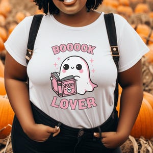 May include: White t-shirt with a pink and black graphic of a ghost reading a book. The text on the shirt reads "BOOOOK LOVER" and "Spooky". The shirt is worn with black overalls, against a background of pumpkins.