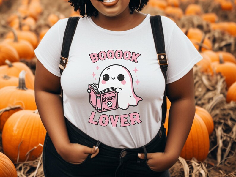 May include: White t-shirt with a pink and black graphic of a ghost reading a book. The text on the shirt reads "BOOOOK LOVER" and "Spooky". The shirt is worn with black overalls, against a background of pumpkins.